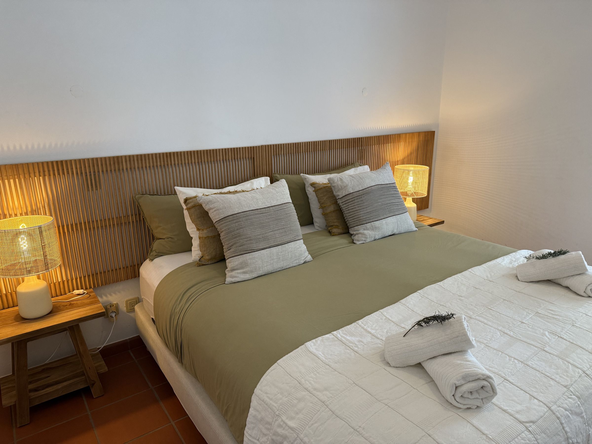 Uma cama de casal com roupa de linho, cabeceira de rattan e candeeiros de cabeceira de luz quente