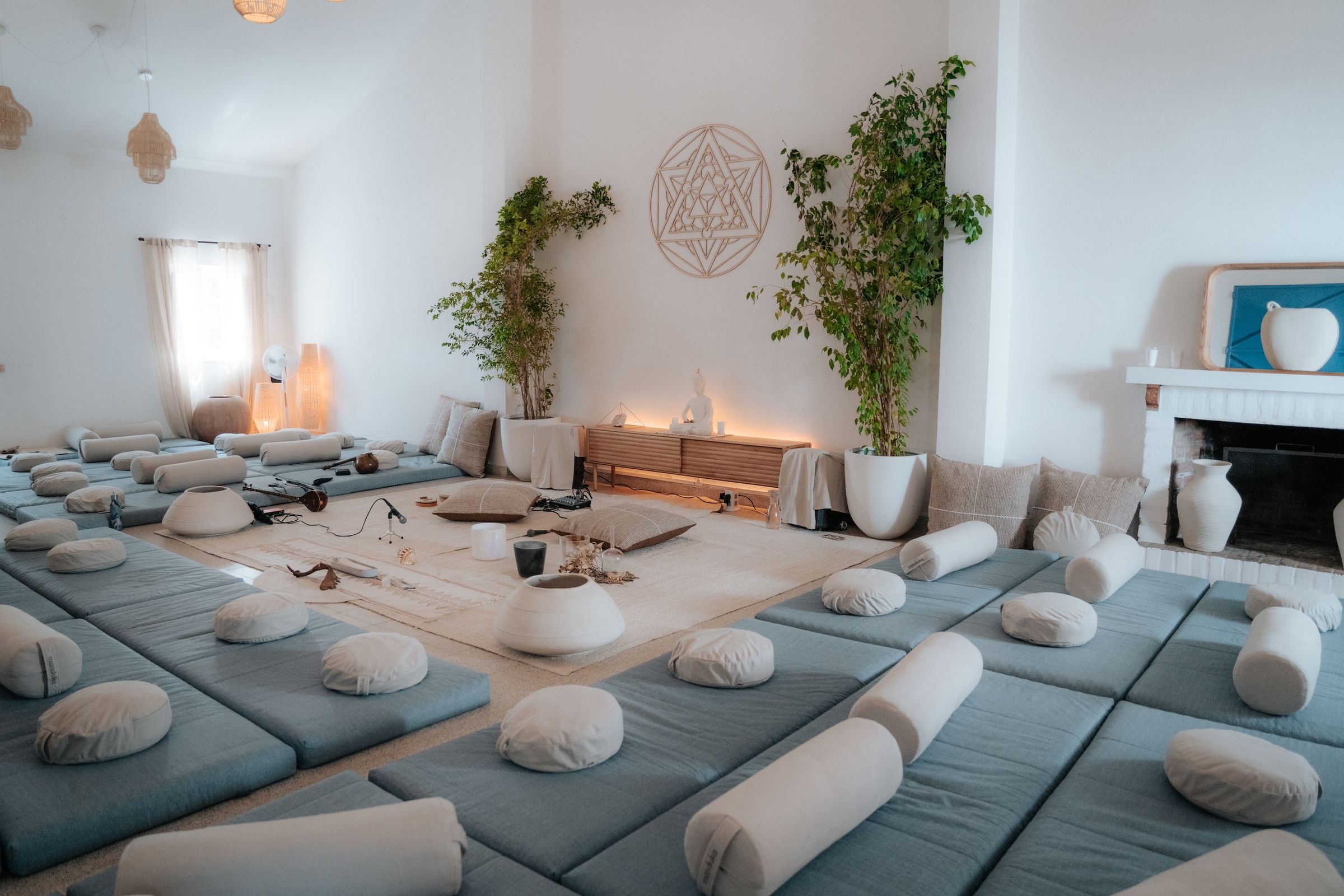 Uma sala branca e luminosa preparada com colchões e almofadas para um retiro, com plantas, uma lareira e um emblema de geometria sagrada