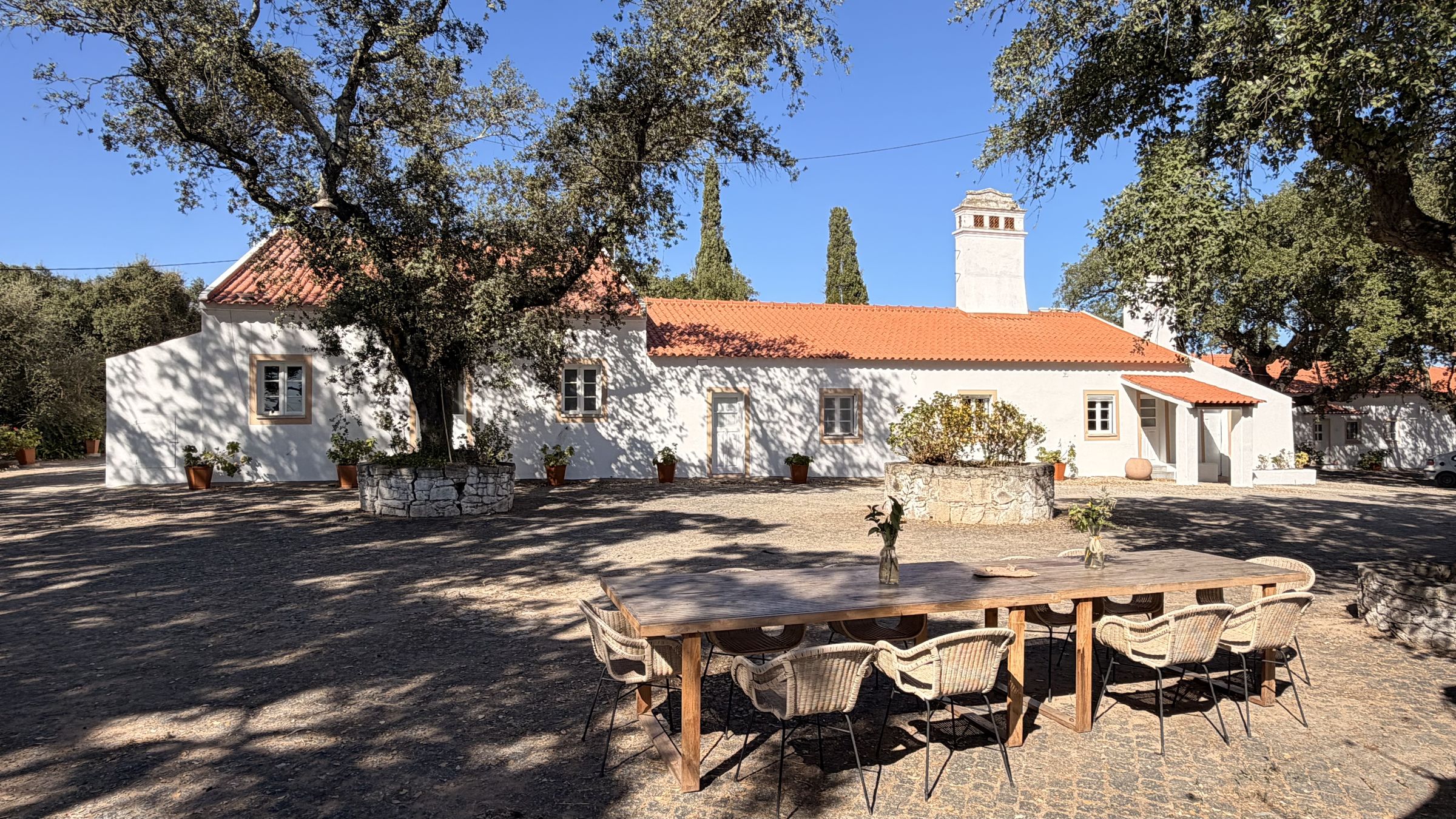 Uma mesa comprida de madeira com cadeiras entrançadas no pátio empedrado, em frente à casa caiada com a sua alta chaminé alentejana