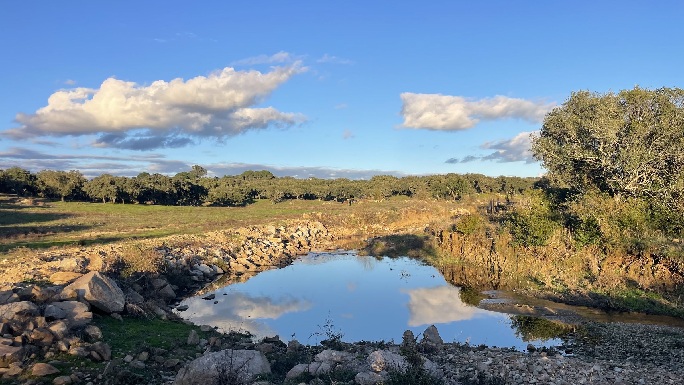 Um pego de água límpida entre rochas de granito a refletir nuvens brancas, com a planície de sobreiros ao fundo