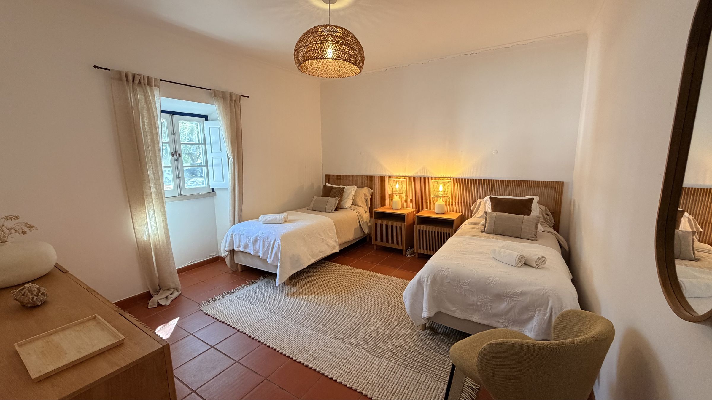 Um quarto de hóspedes com duas camas feitas, cabeceiras de madeira, candeeiros de cabeceira, um tapete de juta e uma janela para o jardim